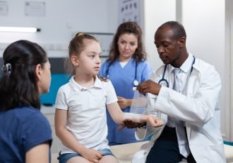 African american pediatrician doctor bandage broken bone of young child arm during medical consultation in hospital office. Girl patient with fractured hand having physiotherapy. Trauma recovery